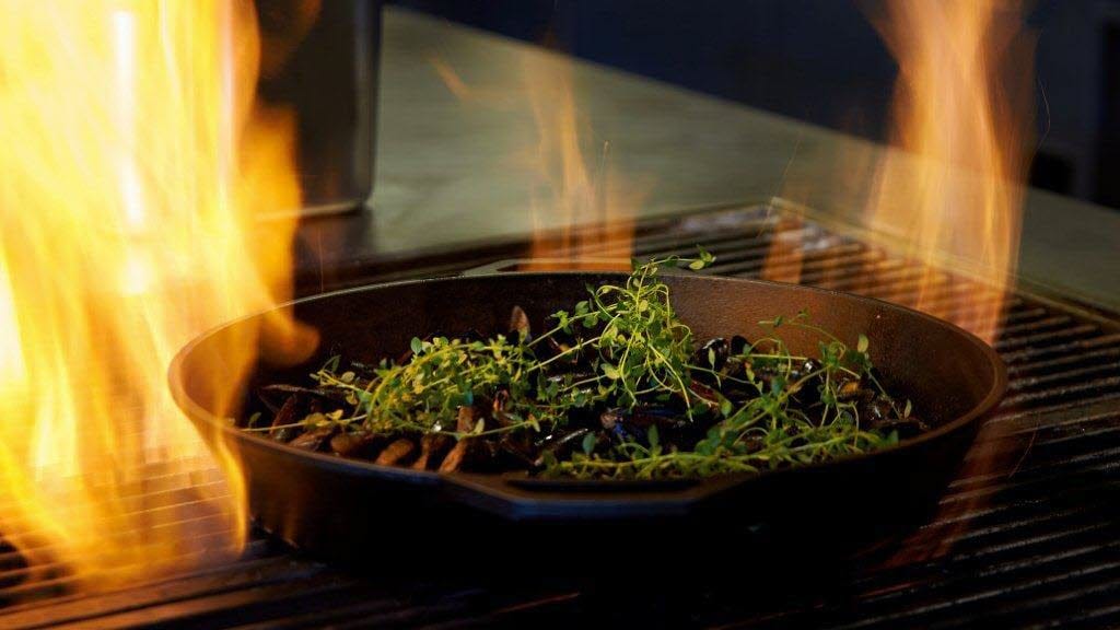 Nærbilde av flammer på grill med stekepanne med krydder og blåskjell på restaurant Ask på Thon Hotel Vettre i Asker.